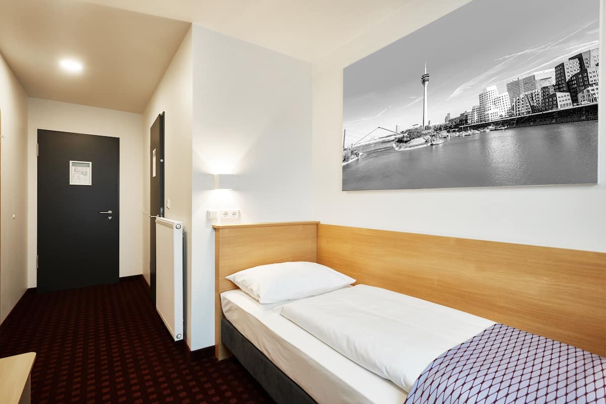 A hotel room with a single bed, a radiator, a door, and a cityscape painting on the wall.