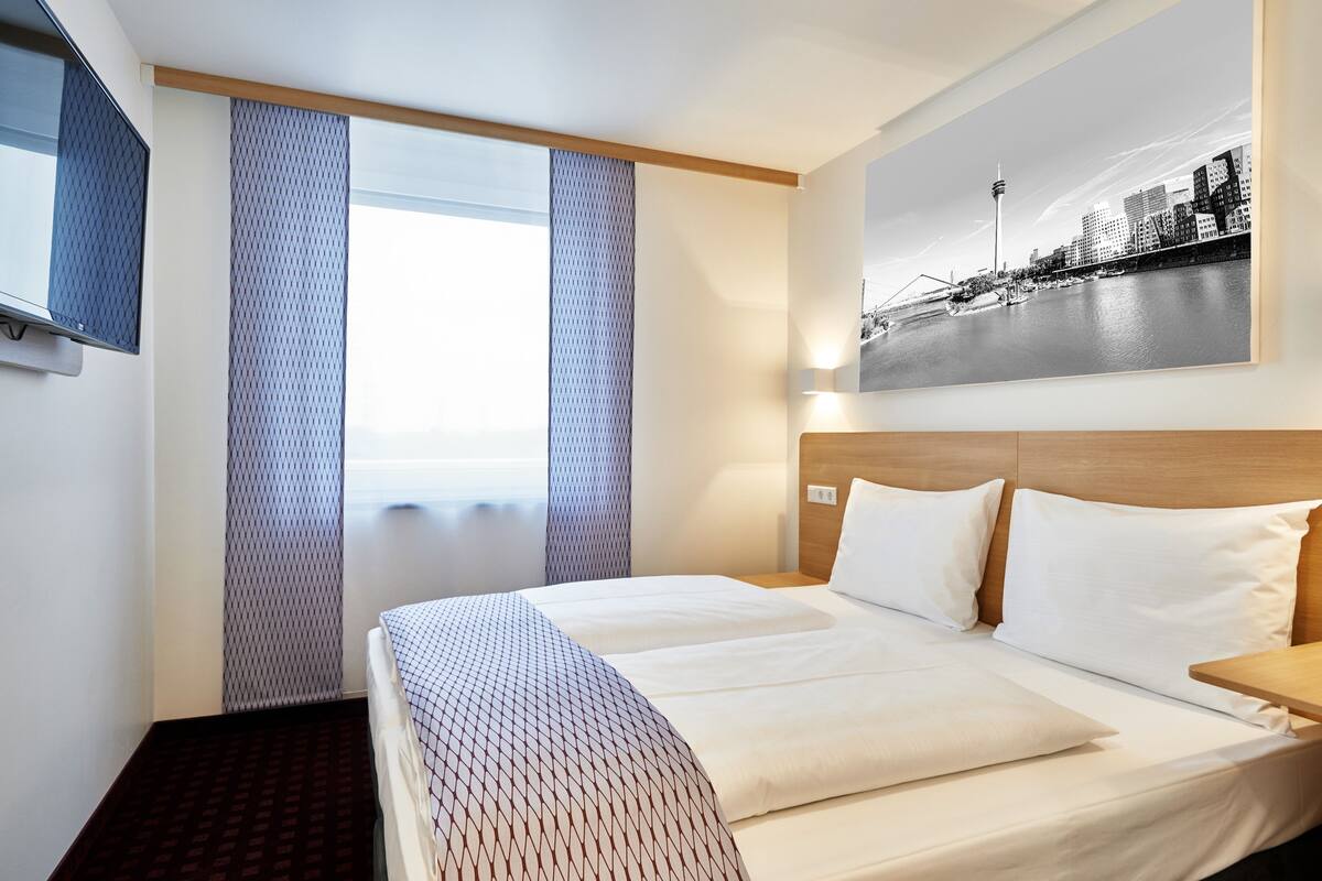 A hotel room with a large bed, a TV mounted on the wall, and a cityscape painting above the bed.