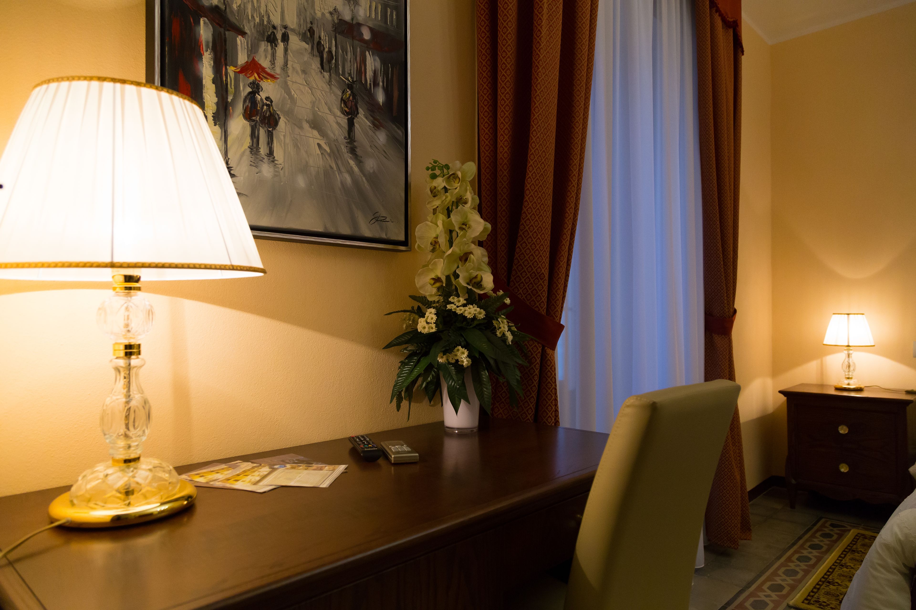 A hotel room with a desk, a lamp, a vase with flowers, a painting on the wall, and a bed with a pillow.
