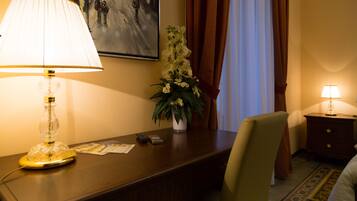 A hotel room with a desk, a lamp, a vase with flowers, a painting on the wall, and a bed with a pillow.