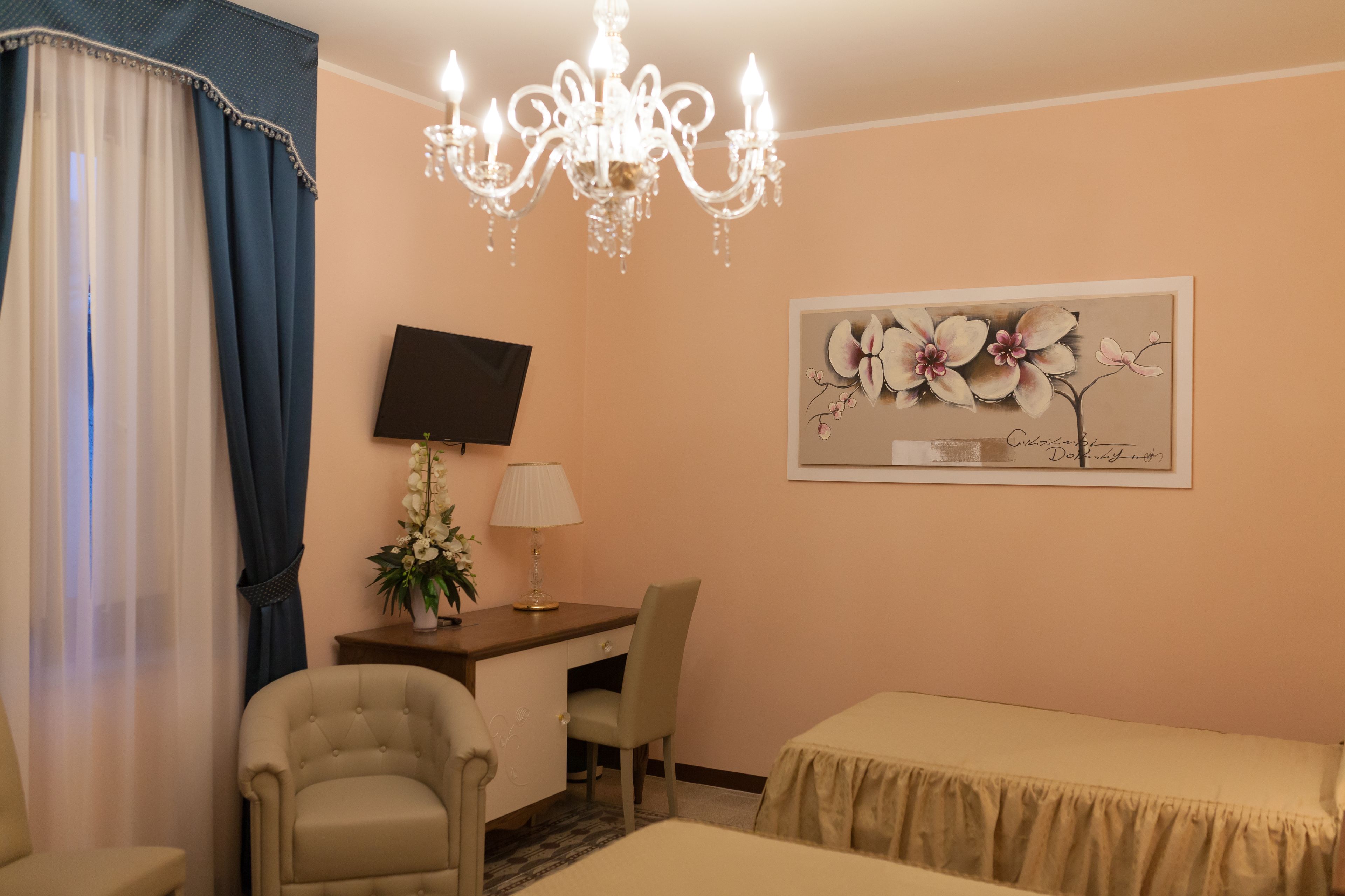 Une chambre d’hôtel équipée d’un bureau, de deux chaises, d’un lit, d’une télévision, d’un lustre et d’un tableau représentant des fleurs.