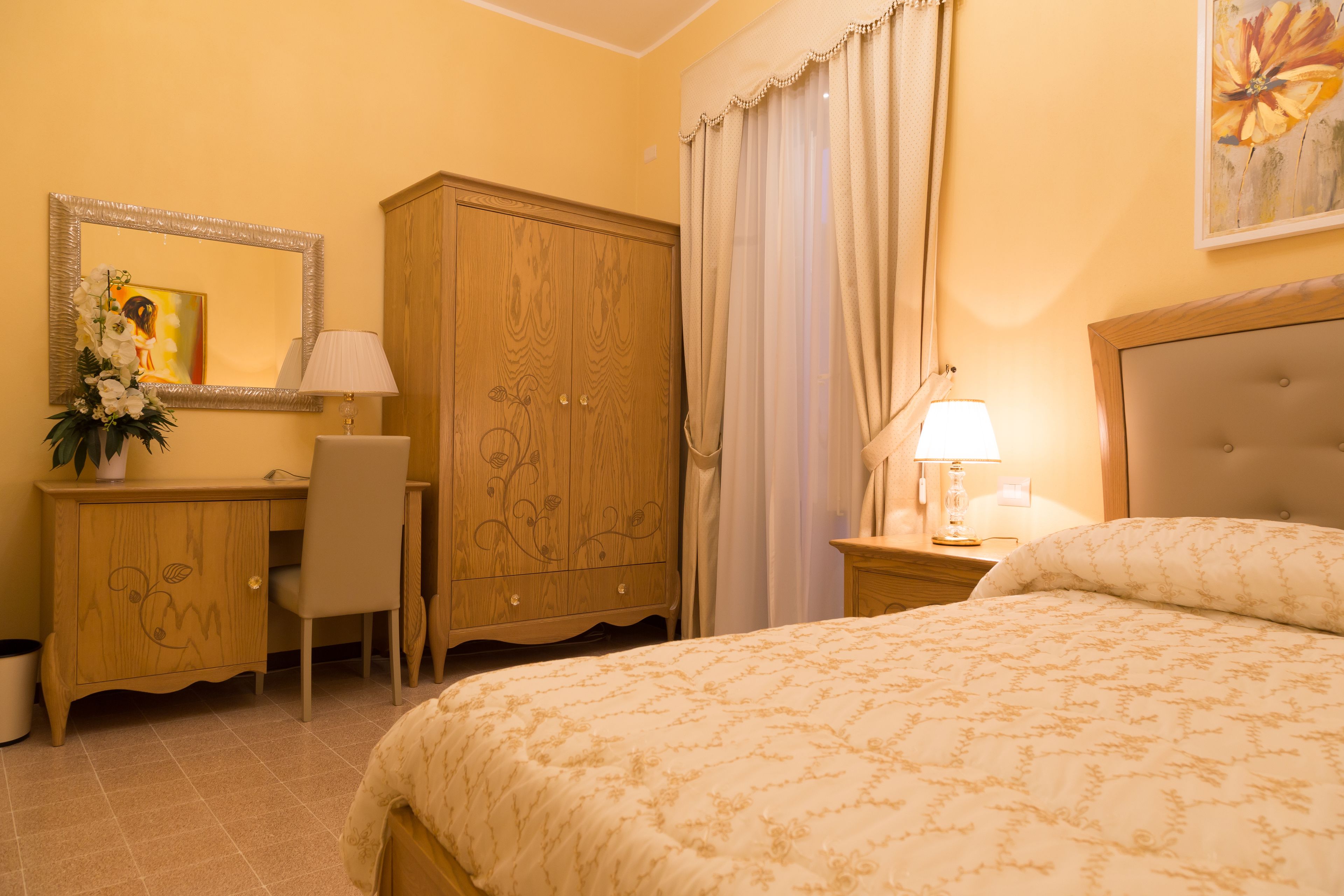 Una habitación de hotel con cama, un armario de madera, un escritorio con silla, una lámpara y un cuadro en la pared.