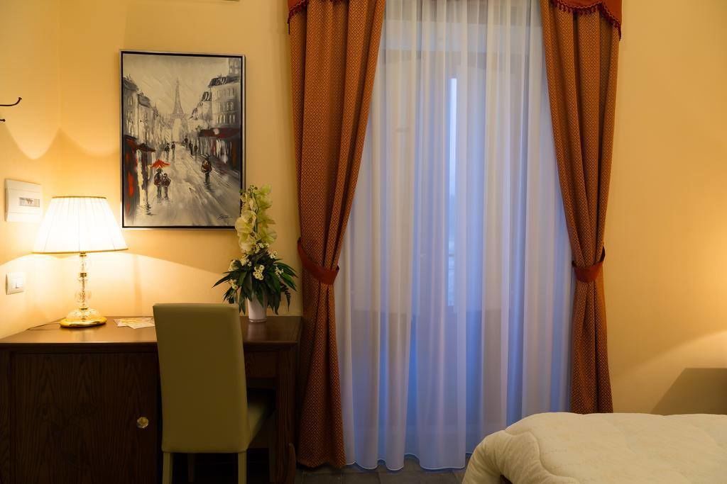 Una camera d'albergo con un letto, una scrivania con una lampada, una sedia, un quadro appeso e una finestra con tende.
