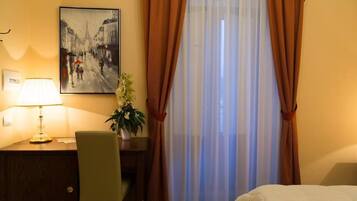 Una camera d'albergo con un letto, una scrivania con una lampada, una sedia, un quadro appeso e una finestra con tende.