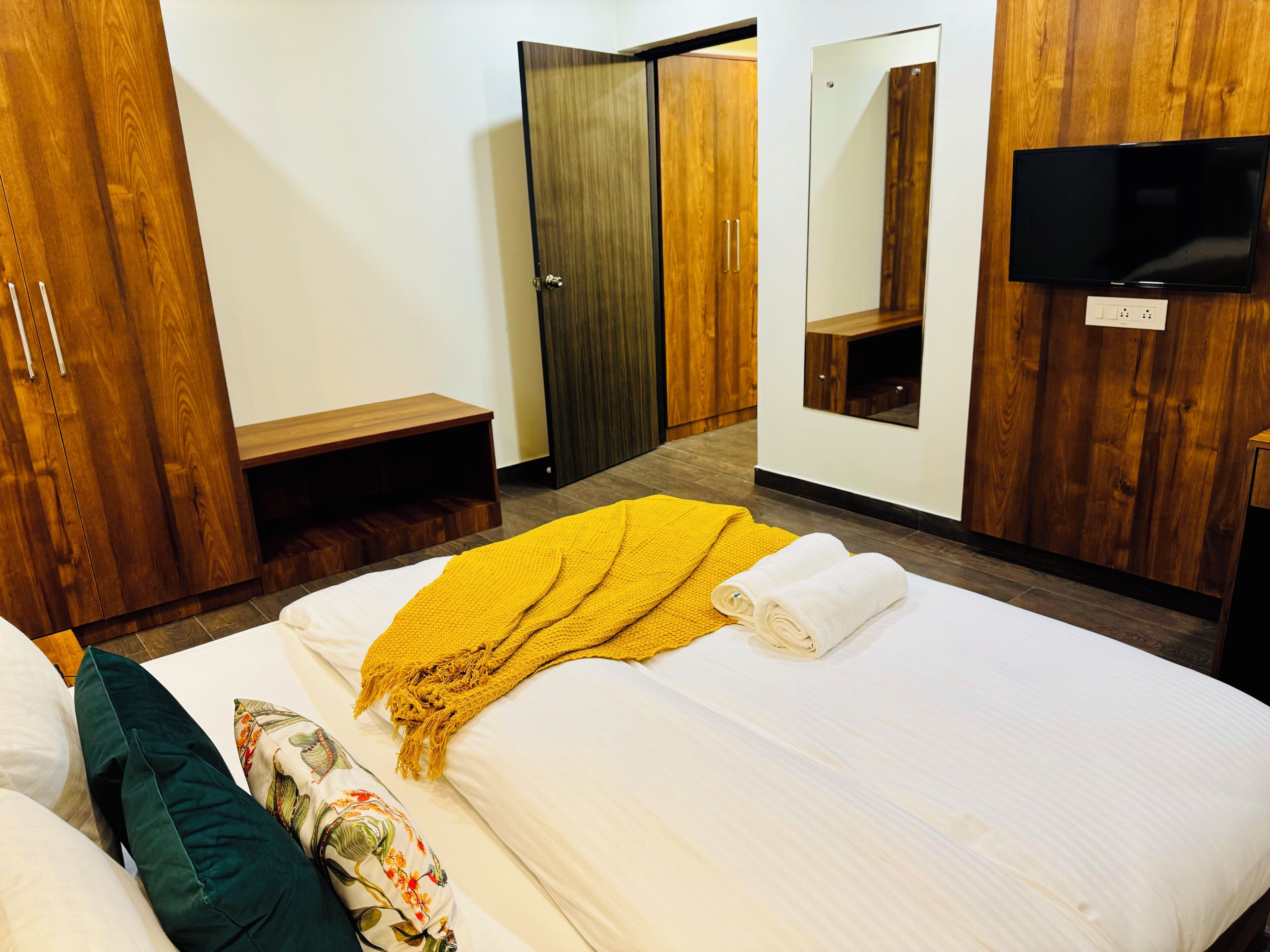A hotel room with a bed, a wooden wardrobe, a small wooden table, a mirror, and a television.