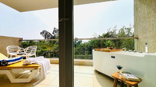 Un balcon avec une table, des chaises, un petit tabouret et un plateau contenant des fruits et une boisson.