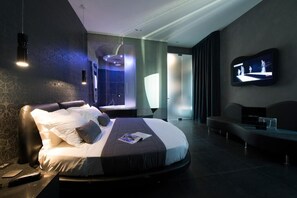 A modern hotel room with a curved bed, a flat-screen TV mounted on the wall, and a seating area with a coffee table.