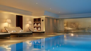 A modern indoor swimming pool with a clear blue water surface, lounge chairs, and a reception area with a reception desk and decorative items.