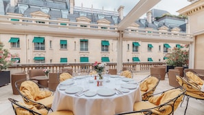 A round table set for dining with wicker chairs, a balcony with a view of buildings, and a railing.