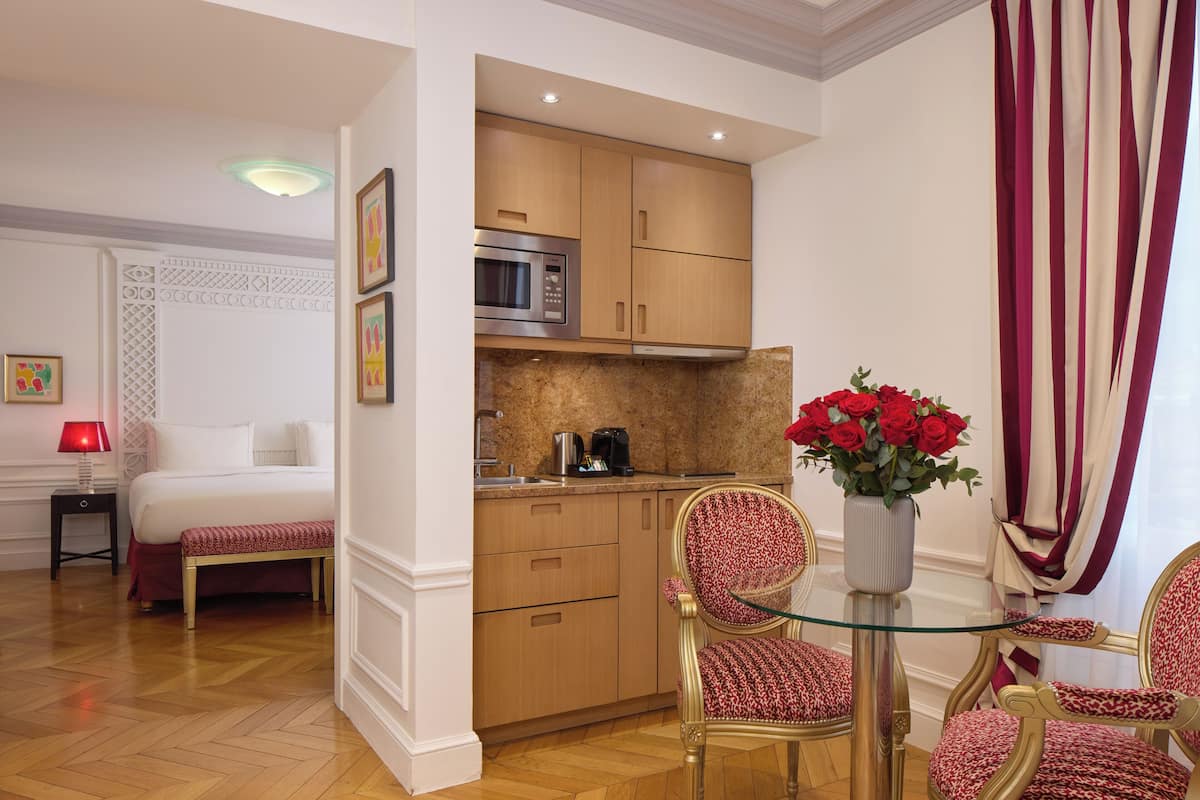 Hotelkamer met een bed, een kleine kitchenette, een zithoek met een tafel en stoelen, en een vaas rode bloemen.