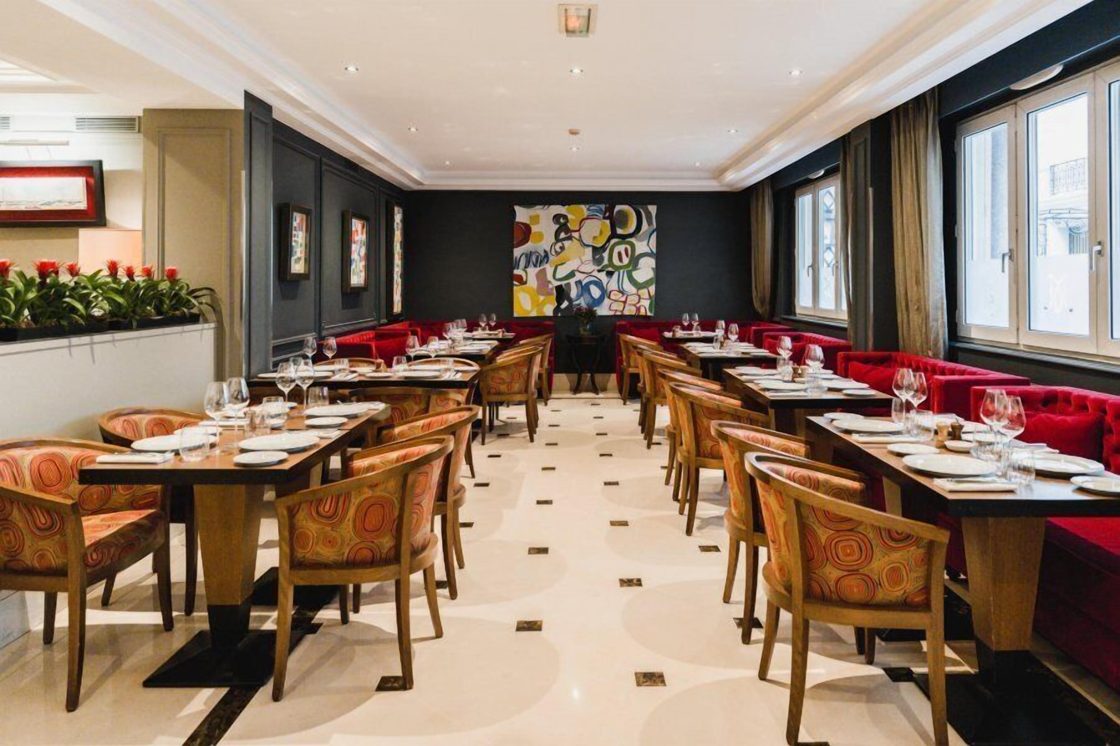 Een restaurant met lange tafels, rode, beklede zitjes en een groot abstract schilderij aan de muur.