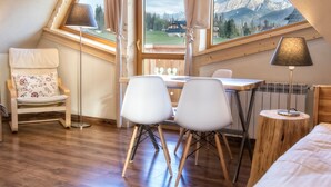 A cozy room with a wooden floor, a dining table with two chairs, a small round table with a lamp, a chair with a floral cushion, and a view of mountains through a large window. - Willa Giewontka (Koscielisko)