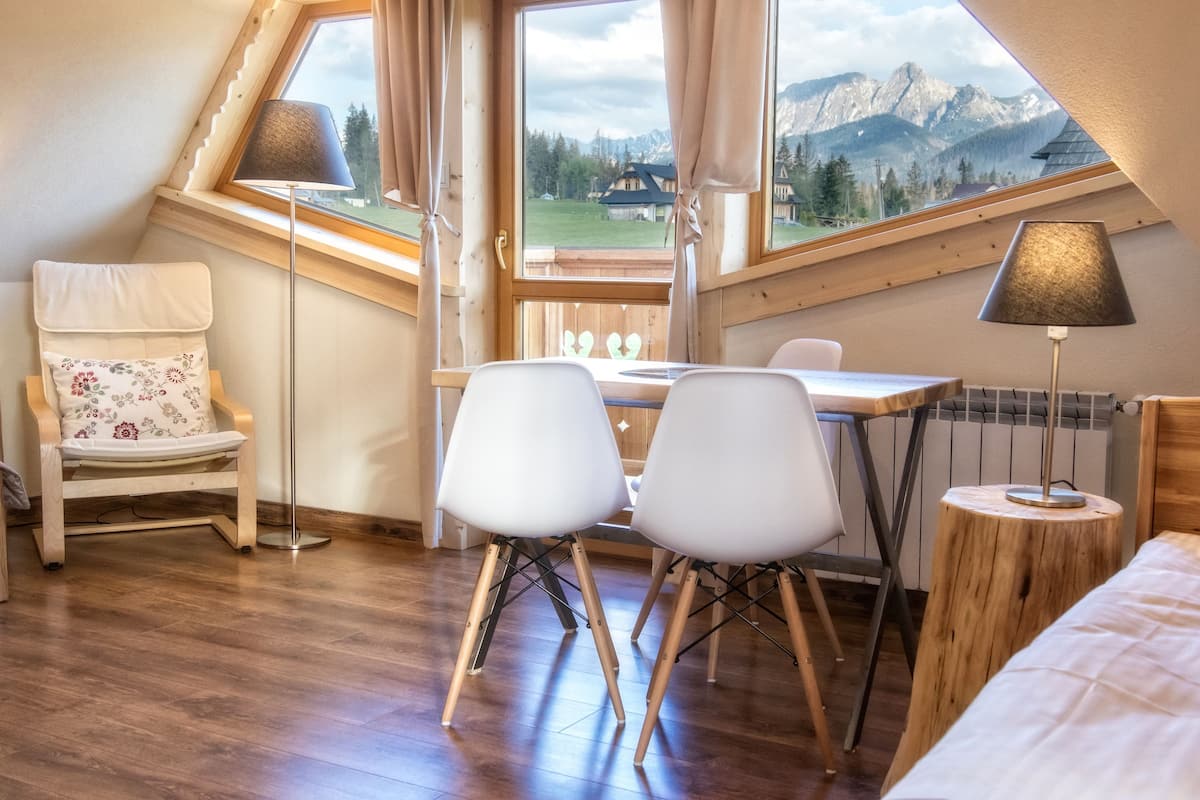 Ein gemütliches Zimmer mit Holzboden, einem Esstisch mit zwei Stühlen, einem kleinen runden Tisch mit einer Lampe, einem Sessel mit floralem Kissen und Blick auf Berge durch ein großes Fenster.