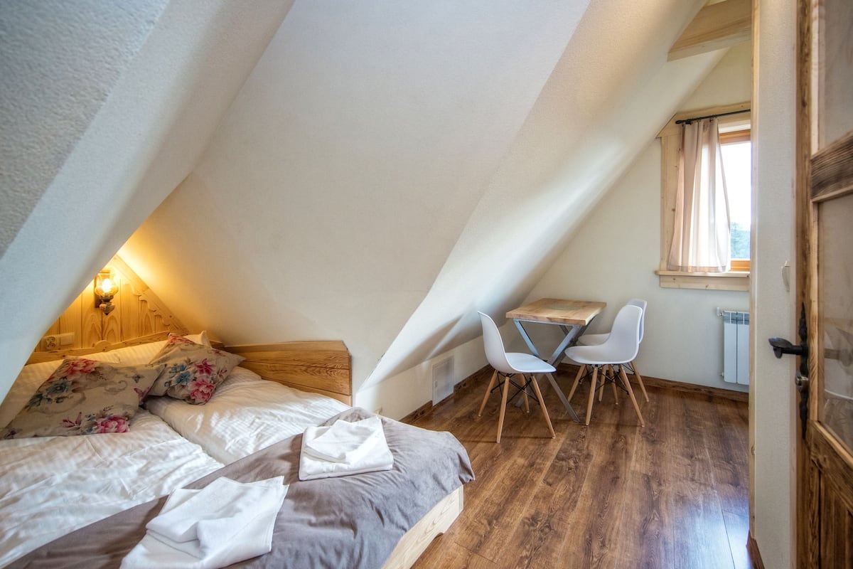 Ein Schlafzimmer mit schrägem Dach, einem Bett mit floralen Kissen, einem Nachttisch mit Lampe, einem kleinen Schreibtisch mit Stuhl und einem Fenster mit Vorhängen.