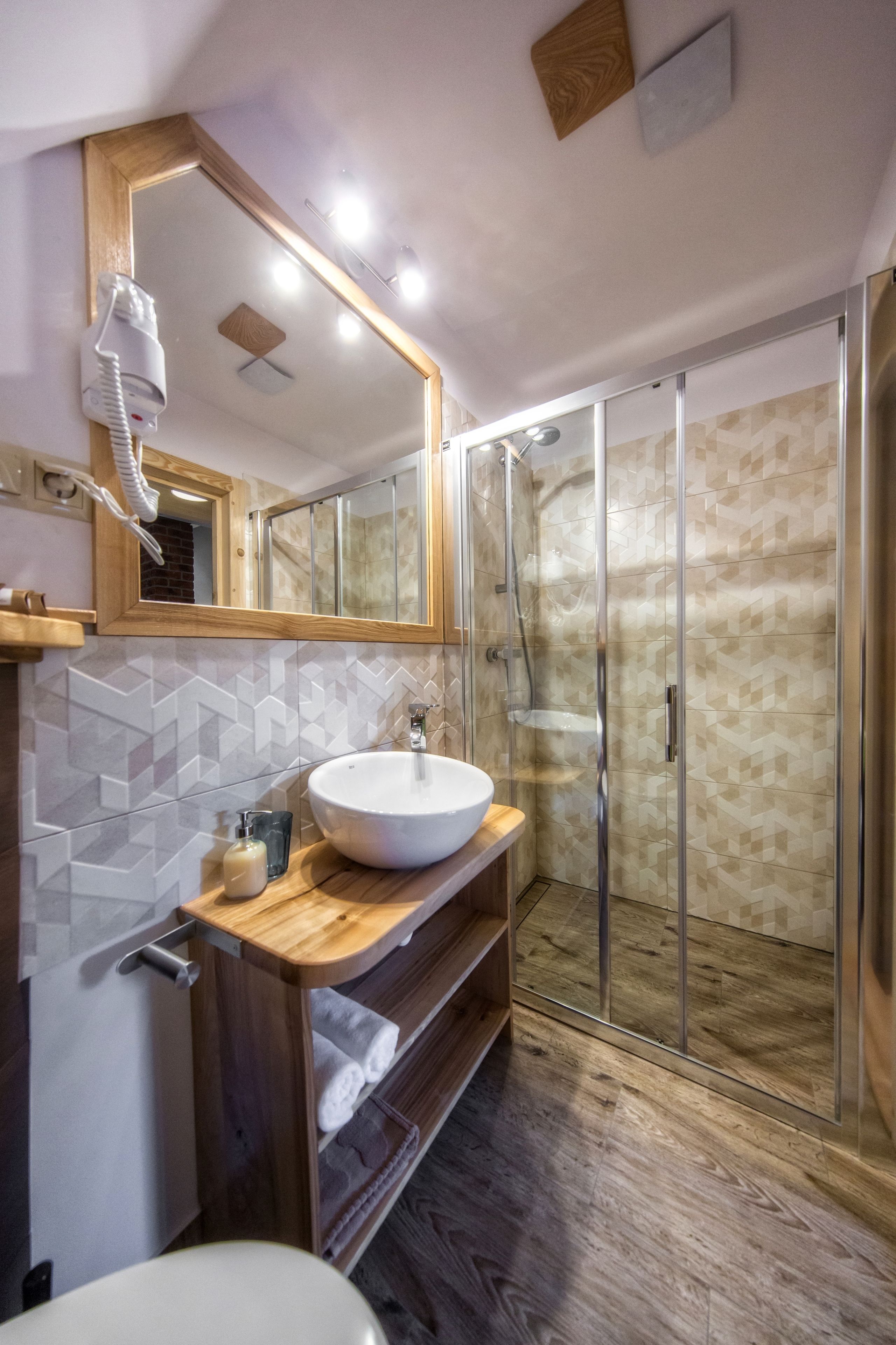 A modern bathroom with a large mirror, a white sink, a glass shower enclosure, and a wooden vanity.