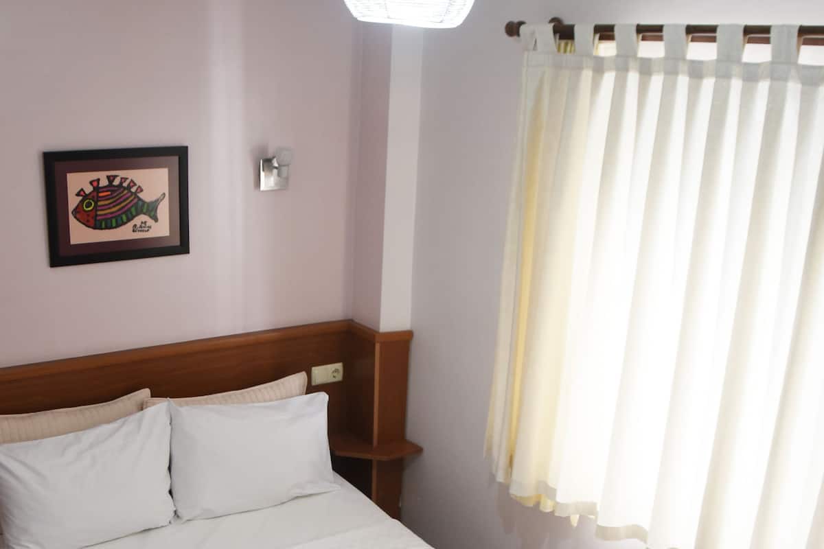 A neatly made bed with white linens, a headboard, a wall-mounted air conditioner, a framed picture, and a window with curtains.