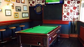 Ein Billardtisch in einem Raum mit gerahmten Bildern an der Wand, einer Dartscheibe und einem Fernseher.