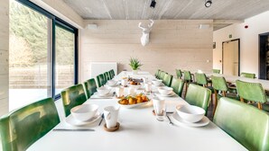 Un coin repas avec une longue table dressée pour le repas, des chaises à l’assise verte et une sculpture représentant une tête de cerf au plafond.