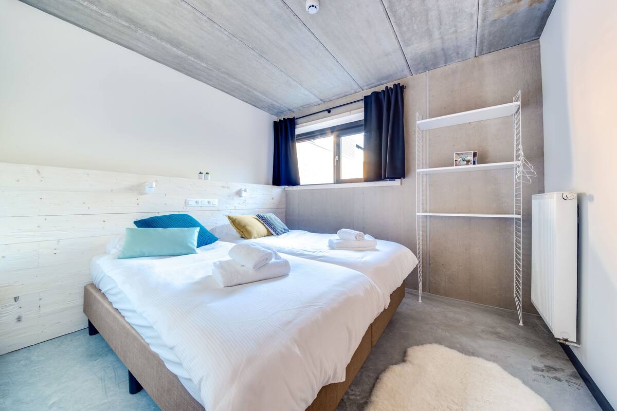 A bedroom with two beds, a white headboard, a window with curtains, a white radiator, and a white shelf unit.
