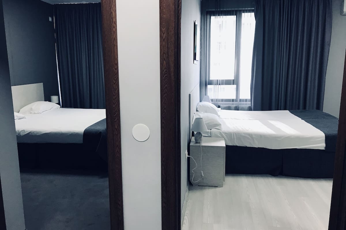 A hotel room with two beds, a bedside table with a lamp, a small trash bin, and a window with curtains.