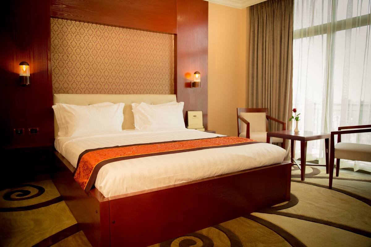A hotel room with a large bed, two bedside tables, a chair, a small table, and a large window with curtains.