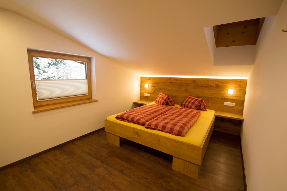 Een slaapkamer met een houten bed, een raam met zonwering en een schuine kap.