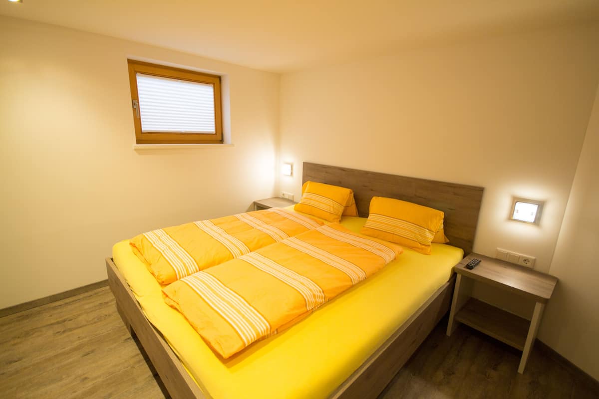 Een netjes opgemaakt bed met een houten hoofdbord en een heldergele spreidek. Er staan twee nachtkastjes met lampen, en er is een raam met zonwering.