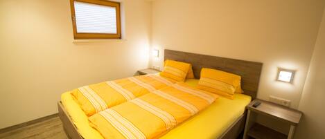 Cama bem arrumada com cabeceira de madeira e colcha amarela vibrante. Há duas mesas de cabeceira com abajures, e uma janela com persianas.