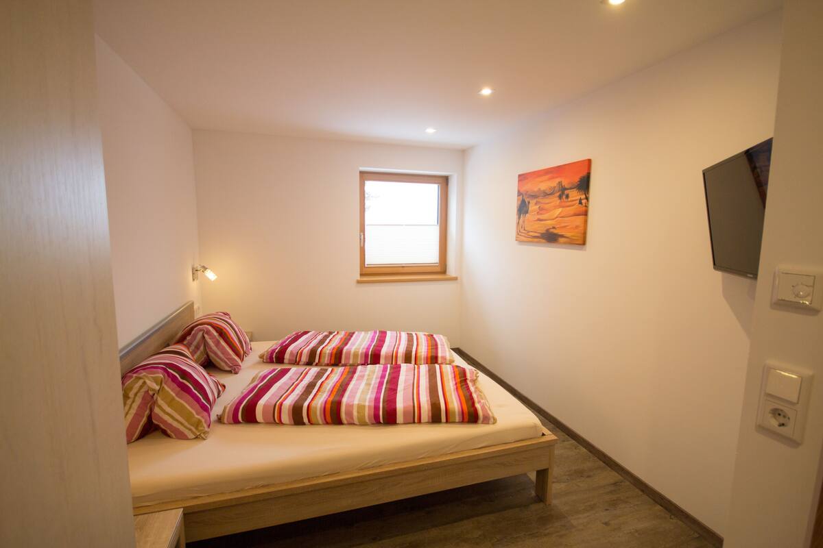 Een kleine, goed verlichte slaapkamer met een enkel bed, een raam en een wandgemonteerde televisie.