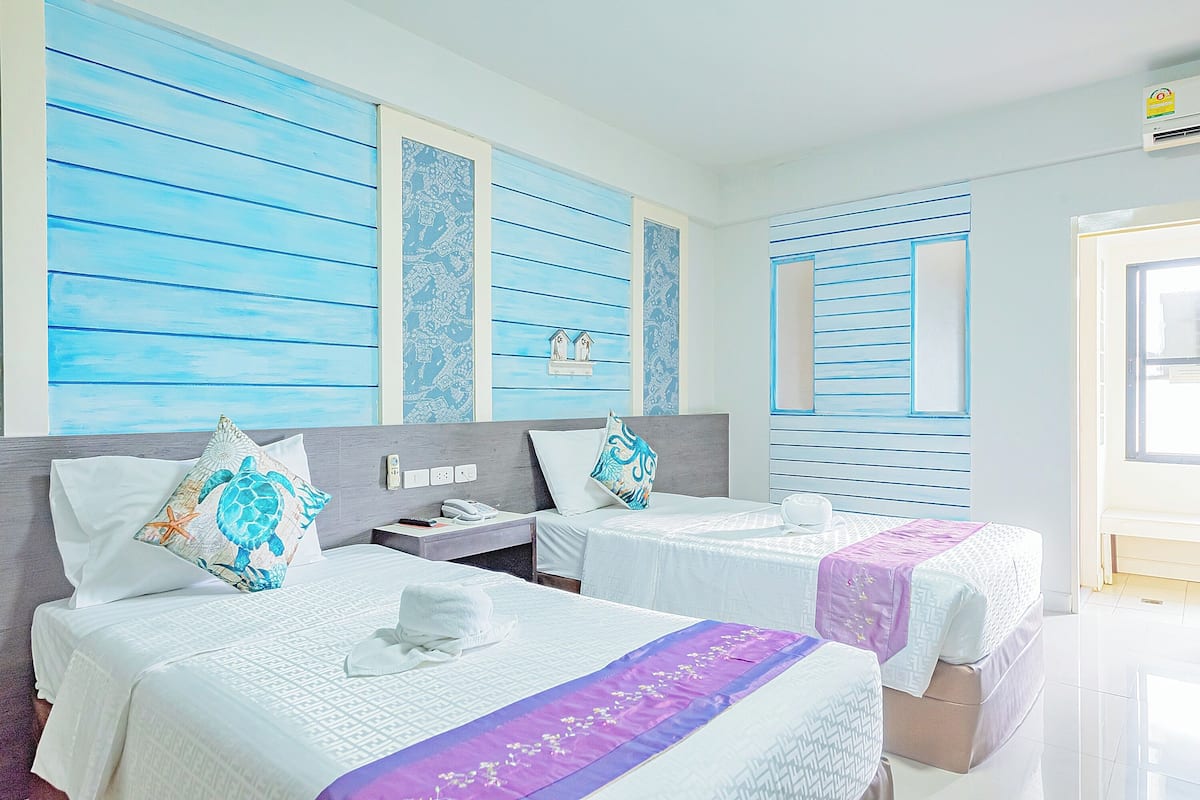 Ein Hotelzimmer mit zwei Betten, jeweils mit Kissen und Handtuch. Ein Nachttisch mit Telefon und einer kleinen Ablagefläche. Die Wände haben ein blau-weiß gestreiftes Muster.