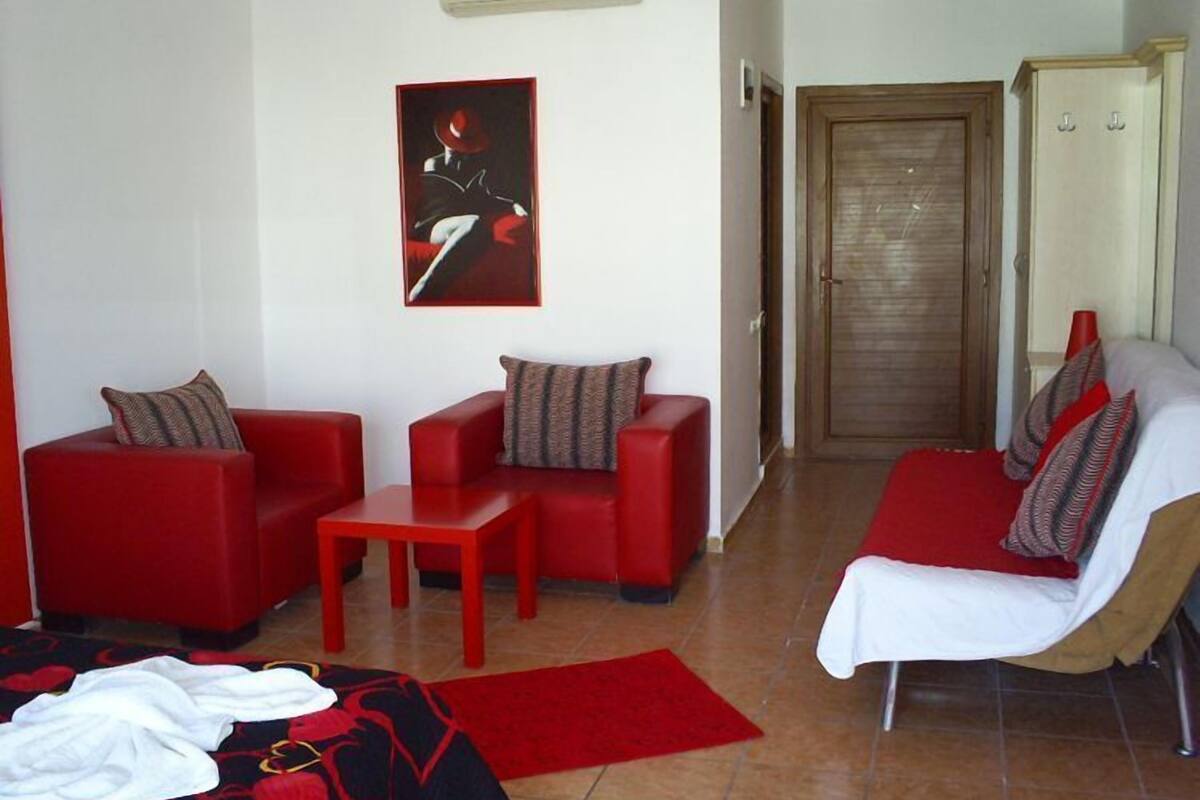 Una sala de estar moderna con sofás rojos, una mesita y una silla blanca. Se ve una puerta al fondo y una alfombra roja en el suelo.