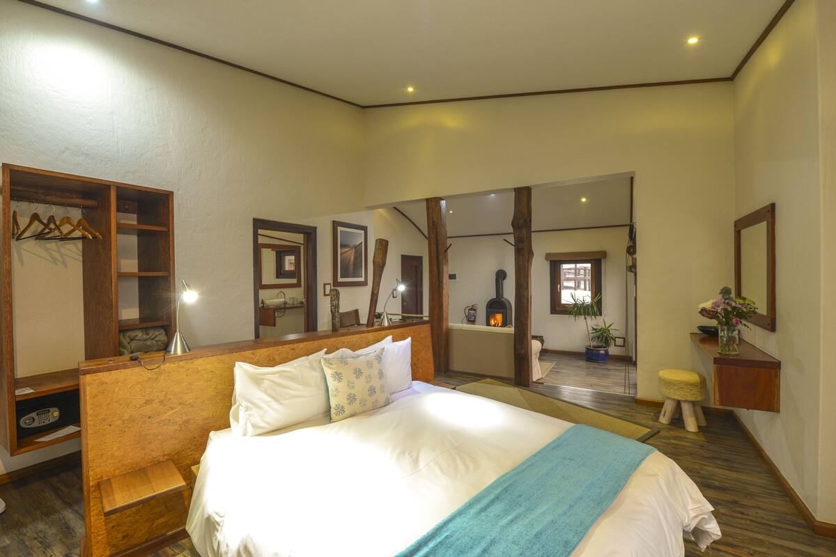 Une chambre avec un grand lit, une tête de lit en bois, des tables de chevet, un miroir et une vue sur une cheminée à travers une porte ouverte.
