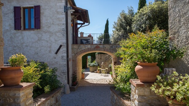 A stone pathway leading to a rustic building with a wooden bridge, potted plants, and outdoor seating.