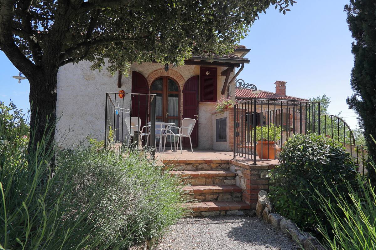 A rustic stone house with a covered patio, white chairs, and a small garden.