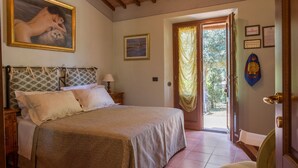 A bedroom with a bed, a nightstand, a lamp, a painting, a door with curtains, and a wall with framed certificates.