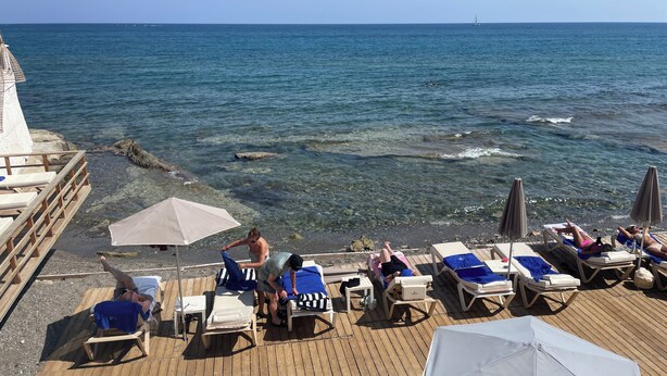 A wooden deck with lounge chairs and umbrellas, overlooking a calm sea with a sailboat in the distance. - Aristo (Hersonissos)