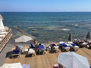 Una terraza de madera con sillones de descanso y sombrillas, con vistas a un mar tranquilo y un velero a lo lejos.