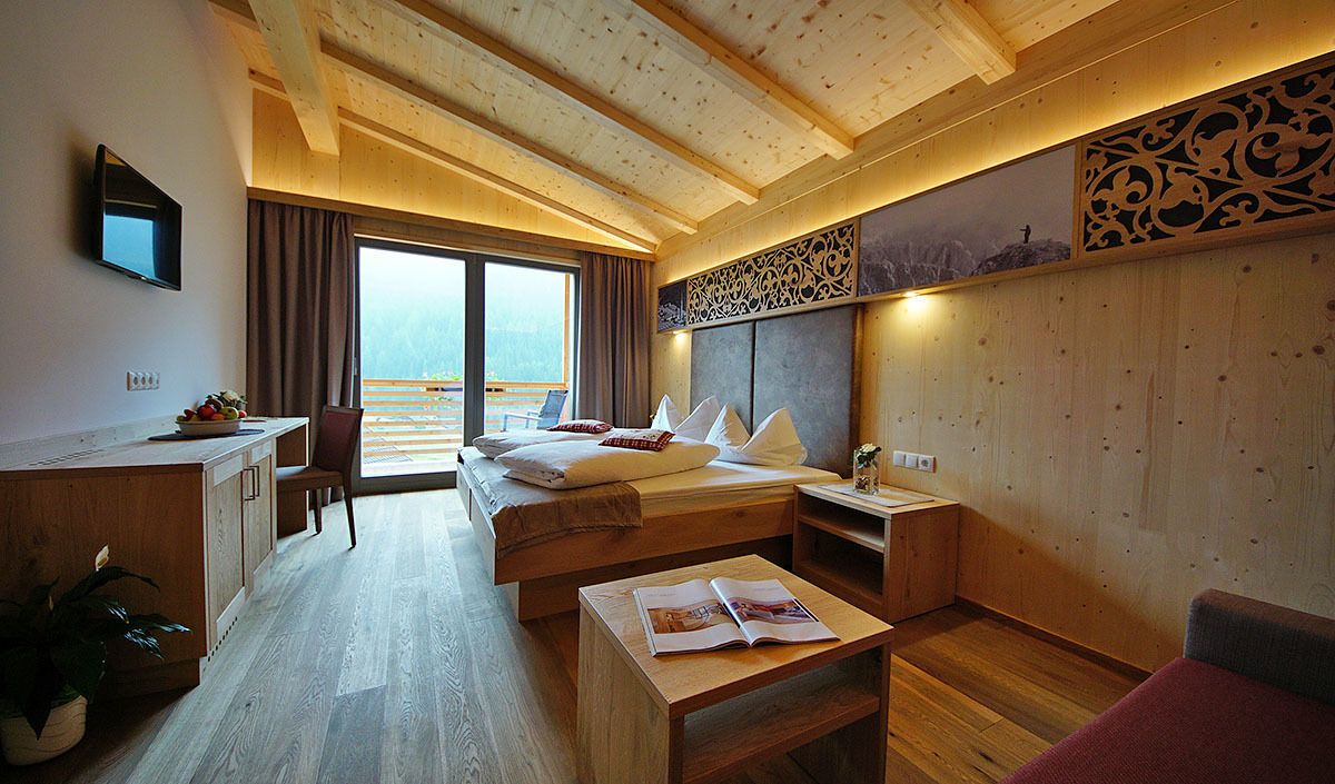 A modern hotel room with a wooden ceiling, a large bed with pillows, a desk with a chair, a small table with a magazine, and a view of the outdoors through a large window.