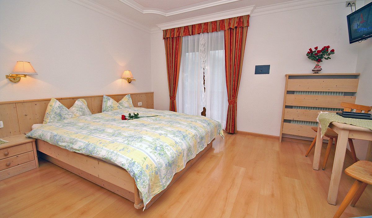 A bedroom with a wooden bed, two bedside lamps, a wooden dresser, and a television on the wall.