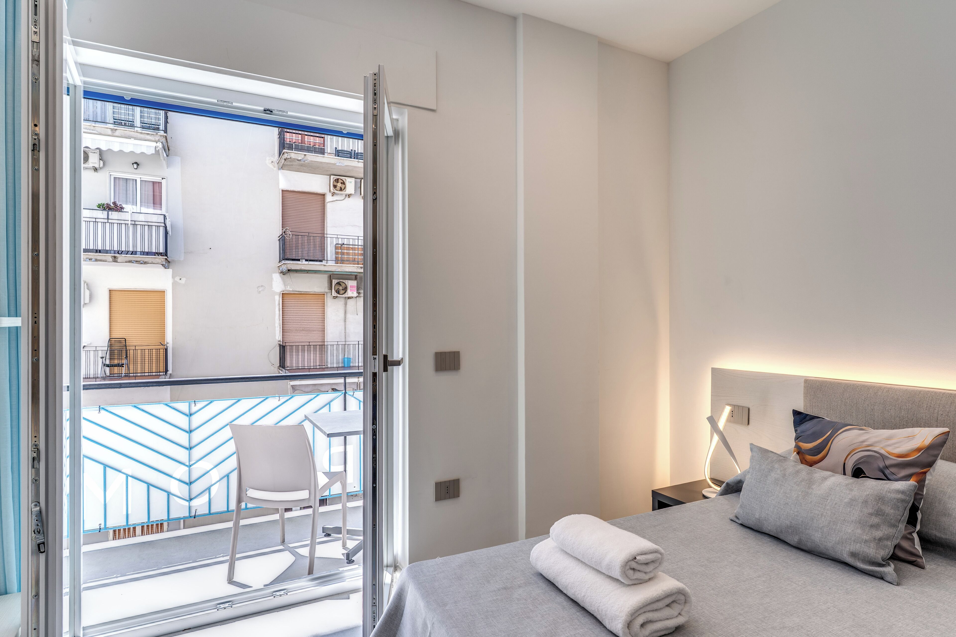 Habitación de hotel con cama, mesita de noche, una silla y vista a un balcón con barandilla y otra silla.