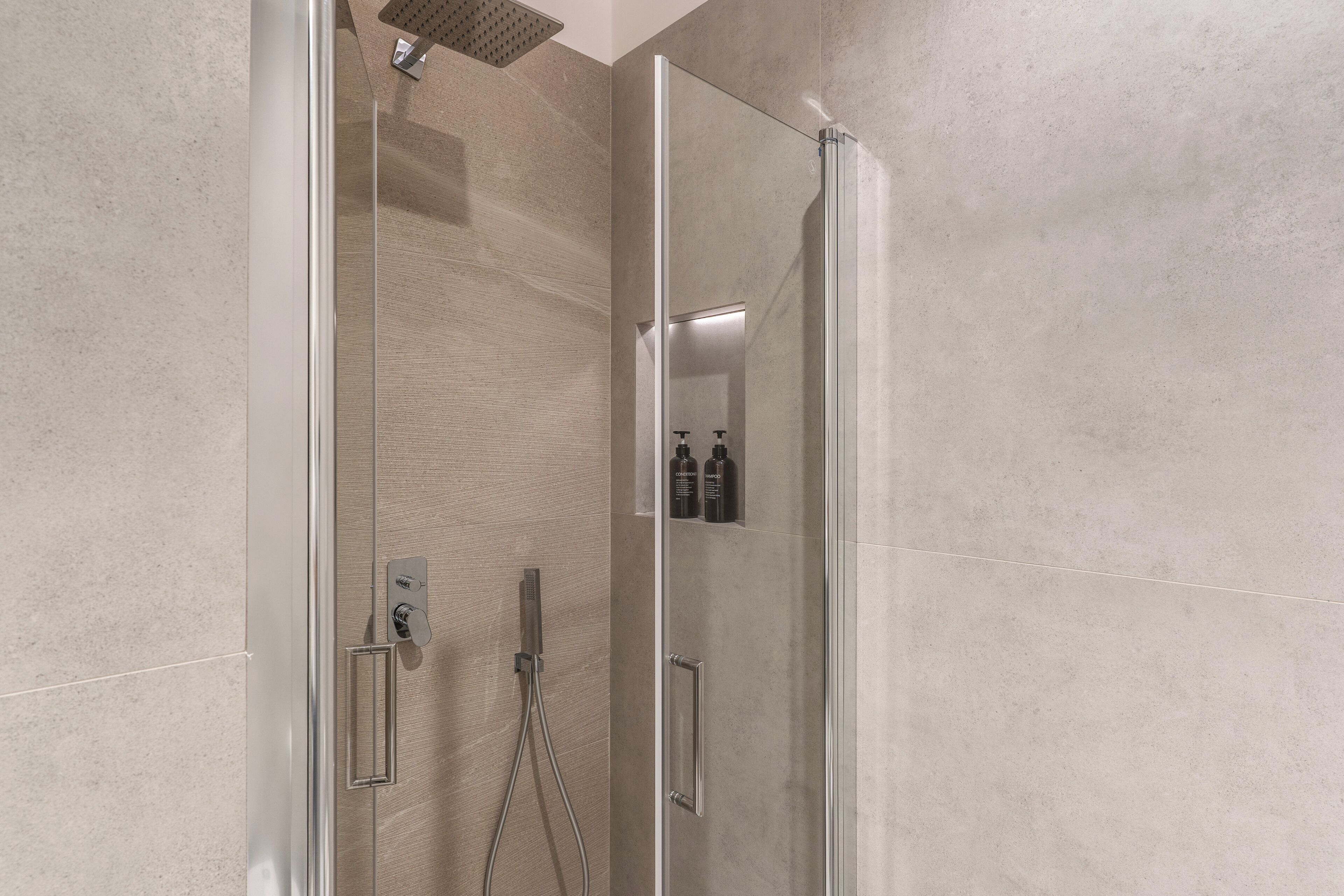 Een moderne douche met glazen wand, handdouche en bedieningspaneel aan de muur.