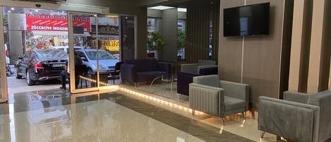 A modern lobby with a polished marble floor, seating area with sofas, a flat-screen TV mounted on the wall, and a visible entrance to a car dealership.