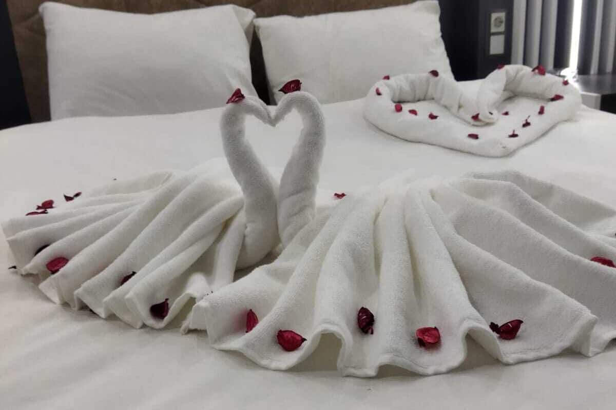 A bed with a white duvet and pillows, a quilted headboard, and a heart-shaped towel decoration made of white towels and red rose petals.