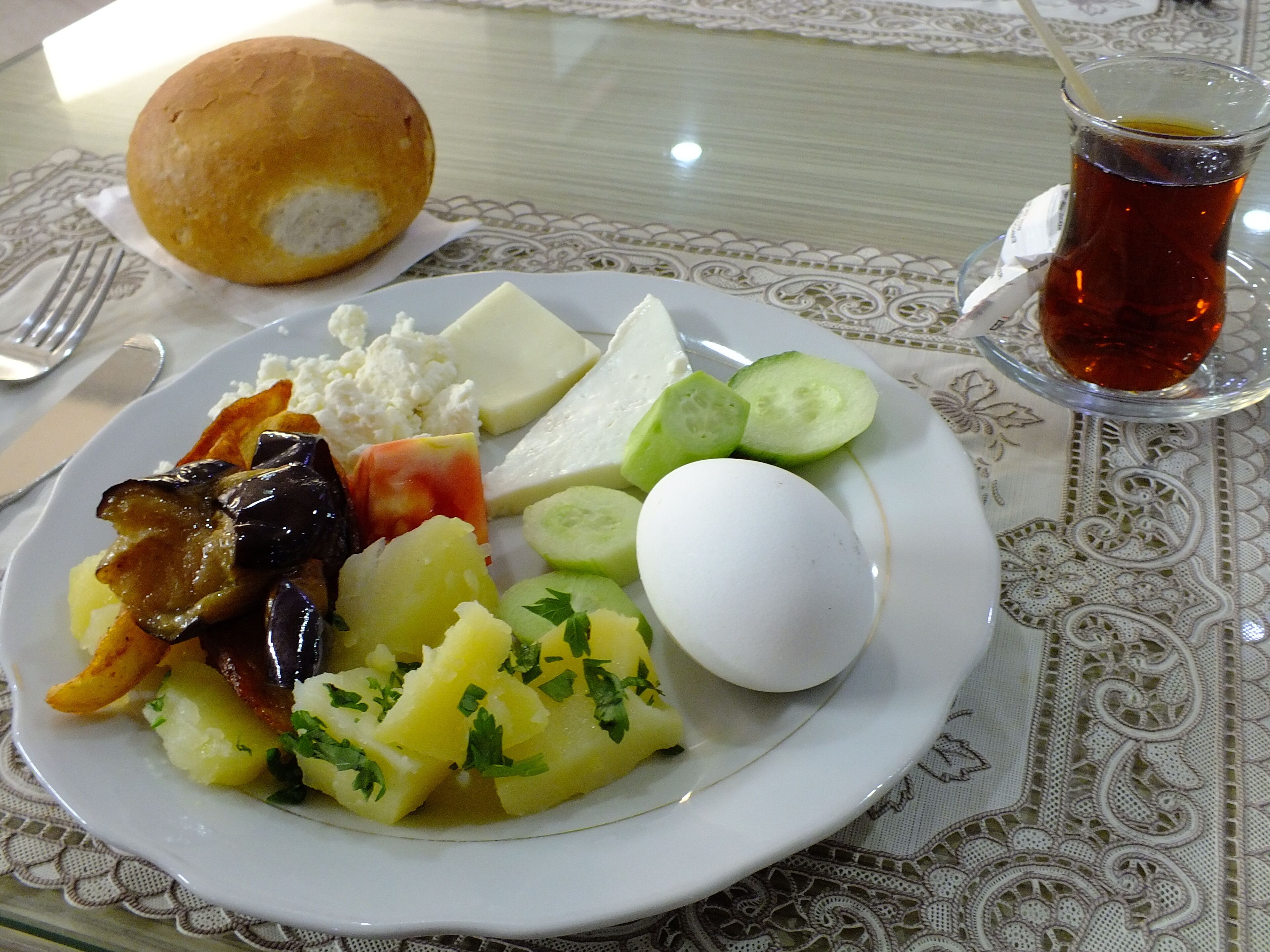A plate of food including a boiled egg, potatoes, cucumbers, cheese, and a roll, with a glass of tea on the table.