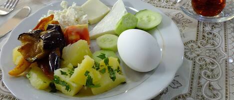 A plate of food including a boiled egg, potatoes, cucumbers, cheese, and a roll, with a glass of tea on the table.