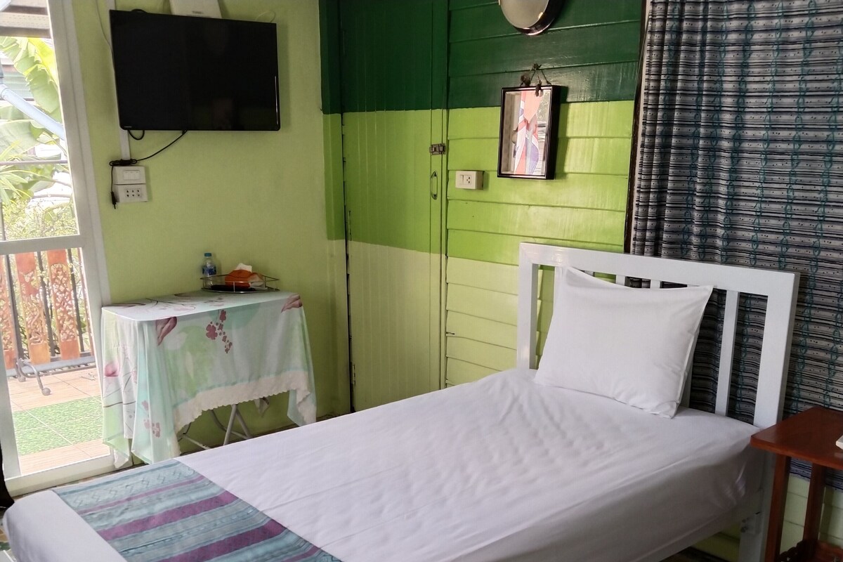 A single-bed room with a green and white color scheme, a table with a floral tablecloth, a television mounted on the wall, and a window with a view of greenery outside.