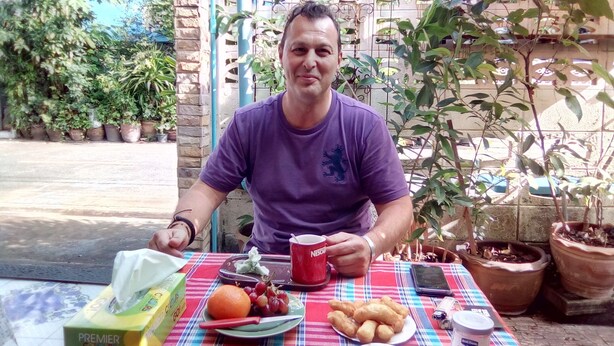 A man sitting at an outdoor table with a meal, surrounded by potted plants and a blue building in the background. - Ban Kru Ae Homestay (Bangkok)