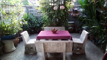 Un patio al aire libre con una mesa y sillas, rodeado de plantas en macetas y una estructura decorativa colgante.