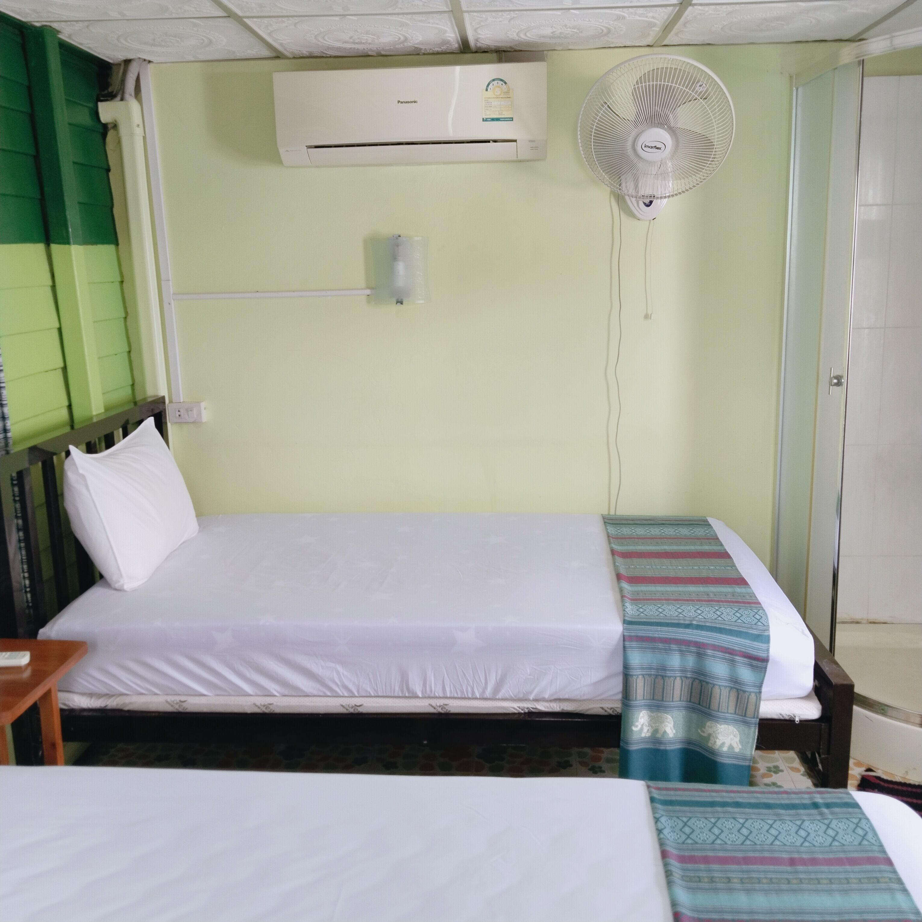 A hotel room with a single bed, a bedside table, an air conditioner, and a wall-mounted fan.