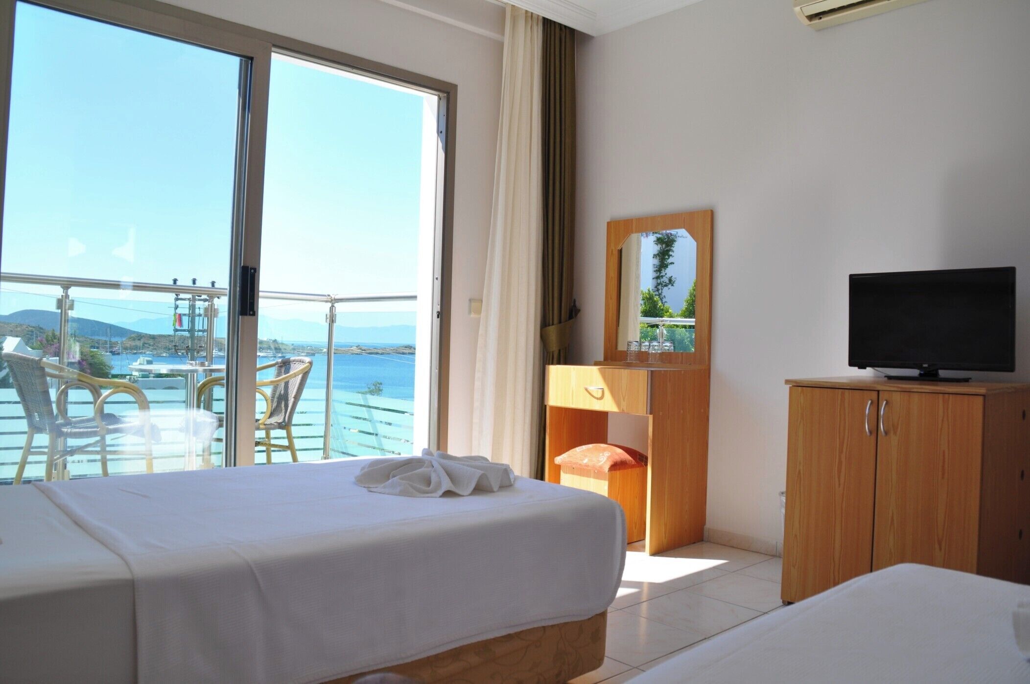 A hotel room with a bed, a television, a wooden cabinet, and a balcony with a view of the sea.